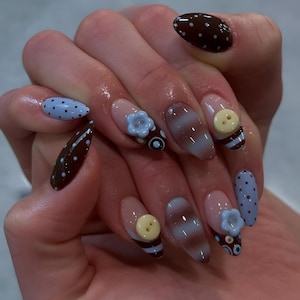 Puede incluir: Una mano con uñas largas y ovaladas con varios diseños. Algunas uñas son marrones con lunares blancos, otras son azul claro con lunares negros. Varias uñas tienen adornos de flores y botones azul claro.