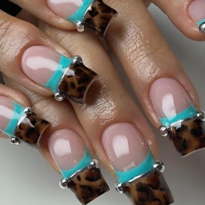May include: Nails featuring a French manicure style with a nude base, a bright turquoise horizontal stripe, and an extended square tip with a brown and black leopard print pattern. Small silver metallic spheres are placed at the corners of the tips.