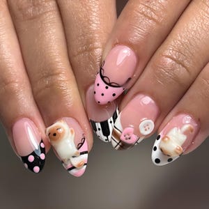 May include: Almond-shaped nails featuring a French manicure base with intricate designs including black polka dots on pink, black and white piano keys, a plaid pattern, a small white button, a pink heart button, and 3D cat figurines.