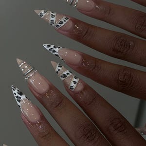 Puede incluir: Primer plano de uñas largas en forma de almendra con base nude. Algunas uñas presentan una punta blanca con diseño de estampado de leopardo negro. Otras tienen anillos plateados y pedrería. Las uñas son brillantes y bien cuidadas.