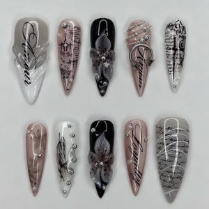 Puede incluir: Un conjunto de diez uñas artificiales largas en forma de almendra. Cada uña presenta diseños únicos, incluyendo detalles florales, escritura y adornos. Los colores incluyen negro, blanco, gris y rosa, con un acabado brillante. Las uñas están dispuestas en dos filas.