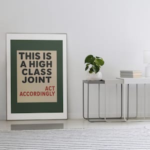 May include: A framed wall art print featuring a dark green border and a beige center panel with black text that reads THIS IS A HIGH CLASS JOINT and red text that reads ACT ACCORDINGLY. The print is displayed in a thin silver frame on a white wall.