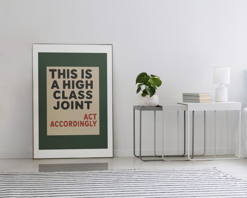 May include: A framed wall art print featuring a dark green border and a beige center panel with black text that reads THIS IS A HIGH CLASS JOINT and red text that reads ACT ACCORDINGLY. The print is displayed in a thin silver frame on a white wall.