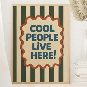 May include: A framed poster with a light beige background and vertical dark green stripes. The poster features the text "COOL PEOPLE LIVE HERE!" in blue, within a red-brown, wavy-edged shape.