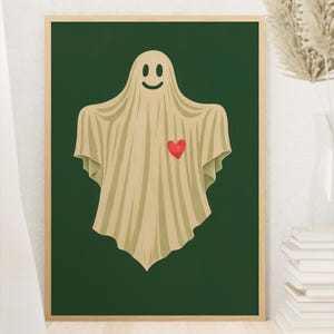 Puede incluir: Una impresión artística enmarcada con una ilustración de un fantasma amigable. El fantasma está cubierto con una sábana beige con un corazón rojo y una cara sonriente sobre un fondo verde oscuro. La obra de arte tiene un estilo minimalista.