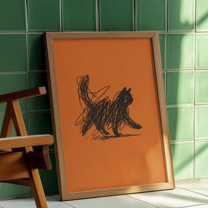 Pode incluir: Uma impressão artística emoldurada com um esboço de gato preto sobre um fundo laranja. O gato é desenhado com linhas rabiscadas, dando-lhe uma aparência lúdica. A moldura é de madeira clara, e a obra está encostada a uma parede de azulejos verdes.