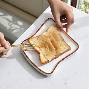Puede incluir: Un plato cuadrado blanco con un borde marrón contiene una rebanada de pan tostado cubierta con rodajas de manzana. Un tenedor dorado está en la esquina inferior izquierda. El plato está sobre una superficie blanca.