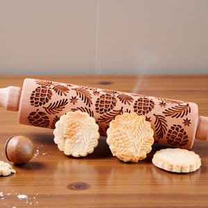Puede incluir: Un rodillo de madera marrón claro con un diseño tallado de piñas y hojas. Se muestran varias galletas en relieve. Las galletas son de color beige claro. La escena está ambientada sobre una superficie de madera.
