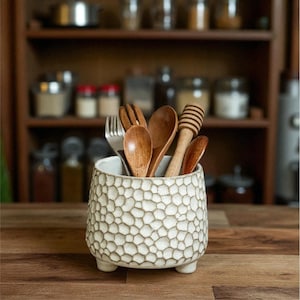 Può includere: Un portautensili in ceramica bianca con un motivo a nido d'ape strutturato, contenente cucchiai di legno, una forchetta e un misurino per il miele. Il portautensili è appoggiato su una superficie di legno, con scaffali della cucina sfocati.