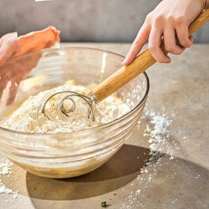 May include: A clear glass bowl filled with flour and a dough whisk. The whisk has a wooden handle and a stainless steel coil. The bowl is on a light brown surface, with flour scattered around it.