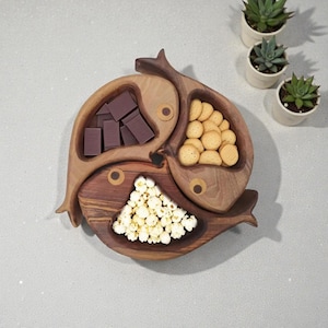 Puede incluir: Una fuente de madera con forma de tres peces, cada uno con diferentes aperitivos: cuadrados de chocolate, galletas y palomitas de maíz. La fuente es de madera marrón clara y oscura, con detalles circulares en los ojos. Suculentas en macetas blancas pequeñas en el fondo.