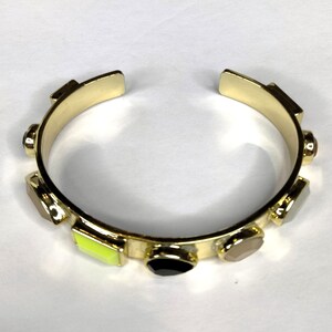 Op de afbeelding: Goudkleurige manchetarmband met kleurrijke edelsteenaccenten. De armband heeft rechthoekige en ovale stenen in zwart, geel, grijs en roze. De armband is aan de bovenkant open en heeft een gladde, gepolijste afwerking.