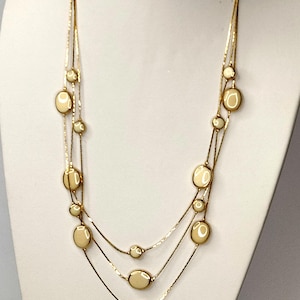 Puede incluir: Un collar dorado de tres hilos con cuentas ovaladas y redondas de color crema. El collar se exhibe en un busto de maniquí blanco. Las cuentas están espaciadas uniformemente a lo largo de los hilos, creando un efecto en capas. El collar es una pieza de joyería clásica.