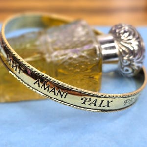 May include: A gold-toned bracelet with engraved text, including the word "PAIX". The bracelet is in front of a perfume bottle with a silver-colored decorative top. The background is a light blue surface.