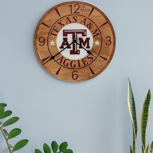May include: A round wooden wall clock with the Texas A&M Aggies logo in maroon and white. The clock face features the words "Texas A&M Aggies" and "12th Man". The clock hands are black.