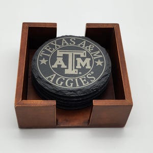May include: A stack of round black slate coasters with rough, natural edges, housed in a square wooden holder. The top coaster features the engraved Texas A&M Aggies logo and text in a circular design, centered on a light gray background.