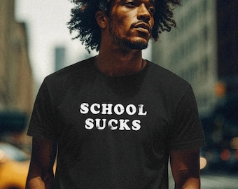 May include: Black t-shirt with the white text "SCHOOL SUCKS". The shirt is worn by a person with dark curly hair, standing outdoors in an urban setting. The person is wearing blue jeans and has their hands in their pockets.