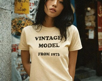 May include: A woman wearing a light tan t-shirt with the text "VINTAGE MODEL FROM 1973" in black. The woman has dark hair and is standing in front of a building with Japanese signage.