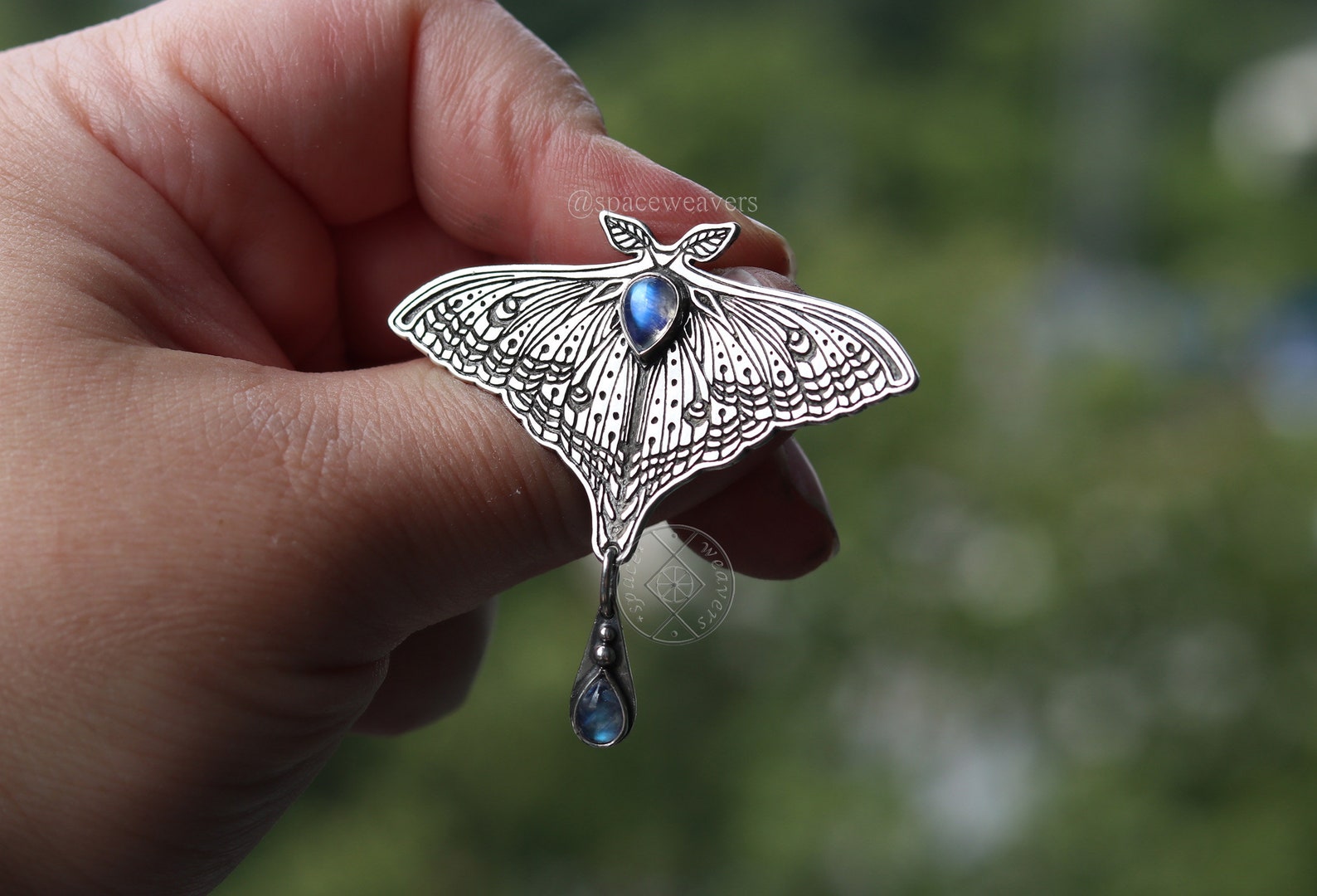 Hand holding a sterling silver luna moth moonstone pendant against a blurred natural background.
