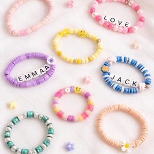 May include: A collection of colorful beaded bracelets. Some bracelets feature names like "EMMAA" and "JACK", while others spell out "LOVE". Smiley face and flower charms add decorative elements. The bracelets are in shades of pink, yellow, purple, blue, and orange.