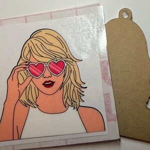 May include: A square sticker featuring a blonde person with shoulder-length hair wearing heart-shaped sunglasses with pink lenses and a white top, positioned next to a brown, die-cut wooden shape with a hole at the top for hanging.