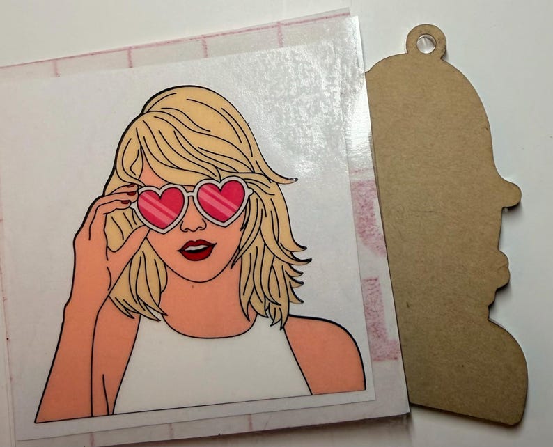 May include: A square sticker featuring a blonde person with shoulder-length hair wearing heart-shaped sunglasses with pink lenses and a white top, positioned next to a brown, die-cut wooden shape with a hole at the top for hanging.