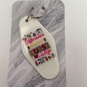 May include: White keychain with the words "Difference maker Nurse wife" and medical-themed graphics. The keychain has a silver key ring and is on a marble-patterned background.