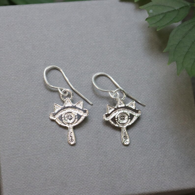 May include: A pair of silver-toned earrings featuring an eye design. Each earring has a detailed eye shape with a crown-like top and a teardrop-shaped bottom. The earrings are suspended from a simple hook.