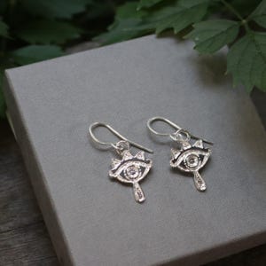 May include: A pair of silver-colored earrings with an eye-shaped design. Each earring features a detailed eye with a crown-like embellishment above and a teardrop-shaped element below. The earrings are displayed on a gray surface.