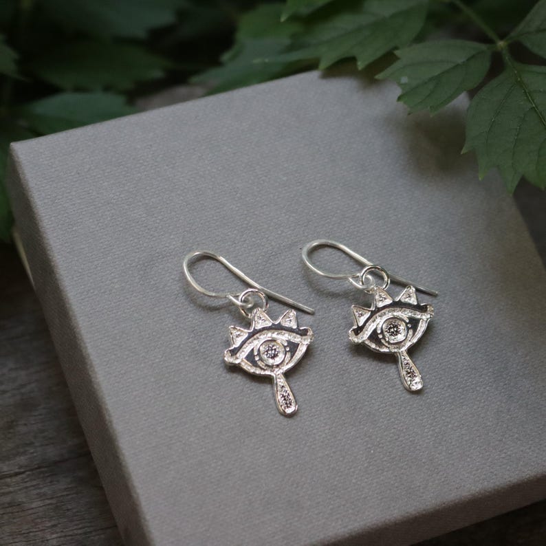 May include: A pair of silver-colored earrings with an eye-shaped design. Each earring features a detailed eye with a crown-like embellishment above and a teardrop-shaped element below. The earrings are displayed on a gray surface.