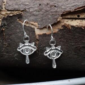 May include: A pair of silver-colored earrings. Each earring features a stylized eye design with a teardrop pendant. The earrings are suspended from a curved hook. The background is a dark brown, textured surface.