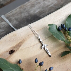 Può includere: Una collana con pendente in argento con un ciondolo a forma di pugnale con dettagli intricati. Il pendente è appeso a una catena delicata. La collana è esposta su una superficie di legno con vegetazione e bacche.