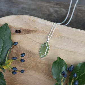 Könnte beinhalten: Eine silberne Halskette mit einem geometrischen, olivgrünen Anhänger. Der Anhänger hat ein facettiertes Design und einen silbernen Rand. Die Halskette wird auf einer Holzoberfläche mit grünen Blättern und blauen Beeren im Hintergrund gezeigt. Die Halskette ist ein Schmuckstück.