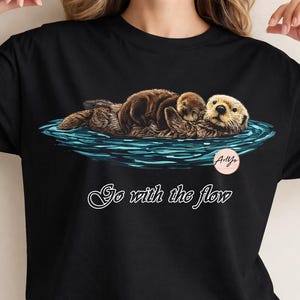 Puede incluir: Camiseta negra con una ilustración de dos nutrias flotando en agua azul. Las nutrias son marrones y beige, una descansando sobre la otra. El texto "Go with the flow" está escrito en cursiva debajo.