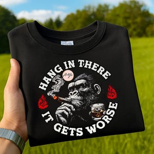 Puede incluir: Sudadera negra de cuello redondo con un gorila fumando un cigarro y bebiendo whisky. El texto dice "Hang in there, it gets worse". Llamas rojas y un círculo rosa acentúan el diseño.