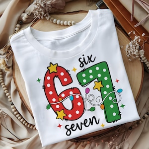 May include: White t-shirt with a festive design featuring the numbers "6" and "7" in red and green, adorned with white polka dots, star accents, and string lights. The word "six" and "seven" are written in black script.