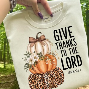 May include: Cream-colored t-shirt featuring a stacked pumpkin design with a leopard print base, orange middle, and white top pumpkin. The design includes floral accents and the text "GIVE THANKS TO THE LORD" with Psalm 136:1.