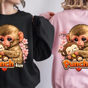 Puede incluir: Sudaderas negras y rosas con un diseño de mono de dibujos animados. La imagen muestra un mono abrazando a un mono más pequeño, rodeado de flores rosas. También se incluye el texto "Punch-kun" y un corazón. Cuello redondo.
