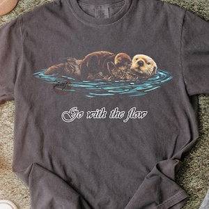 Puede incluir: Camiseta gris oscuro con una ilustración de dos nutrias marinas flotando en agua azul. El texto "Go with the flow" está escrito en blanco debajo. La camiseta está hecha de un material suave.