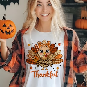 Könnte beinhalten: Weißes T-Shirt mit einem Cartoon-Truthahn-Design, mit einer Schleife, gemusterten Federn und dem Wort "Thankful" in Orange. Die Person trägt ein orange-schwarz kariertes Hemd und hält einen geschnitzten Kürbis.