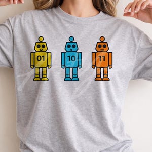 Może przedstawiać: Jasnoszara koszulka z trzema kolorowymi robotami. Każdy robot jest w innym kolorze: żółtym, niebieskim i pomarańczowym, z numerami 01, 10 i 11. Roboty mają czarne obrysy.