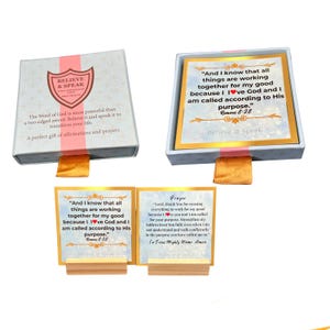 Op de afbeelding: Een geschenkset met affirmatiekaarten en een doos. De doos is grijs met een roze lint en de woorden "BELIEVE & SPEAK". De kaarten hebben gouden randen en inspirerende citaten. Eén kaart luidt: "En wij weten dat alle dingen meewerken ten goede..."