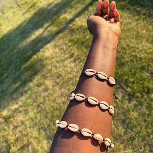 May include: Three bracelets made of small, cream-colored seashells strung on black cords. The bracelets are worn on an arm, with the shells arranged in a row. The background is a grassy area with sunlight.