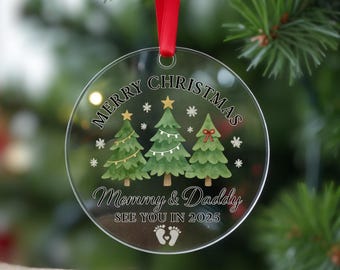 Può includere: Un ornamento natalizio rotondo e trasparente con un nastro rosso. Presenta tre alberi di Natale verdi e la scritta "MERRY CHRISTMAS Mommy & Daddy SEE YOU IN 2025" in nero.