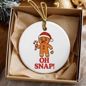 May include: A white ceramic Christmas ornament featuring a distressed gingerbread man wearing a Santa hat and holding a candy cane. The text "OH SNAP!" is printed in red. The ornament is in a brown box with gold string.
