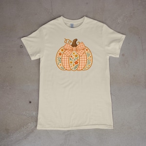 May include: A cream-colored short-sleeve t-shirt featuring a central graphic of a pumpkin. The pumpkin has a light orange gingham pattern, a brown stem, and a large bow, decorated with small orange and yellow flowers and green leaves.