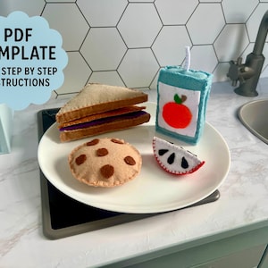 May include: A felt play food set on a white plate. Includes a sandwich, cookie, juice box, and a slice of watermelon. The juice box has an apple applique. The cookie has brown spots. The image also includes the text "PDF TEMPLATE AND STEP BY STEP INSTRUCTIONS".