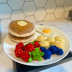 May include: A white plate displays a felt breakfast scene. It includes a stack of pancakes with a butter pat, a fried egg, muffins, red strawberries, blueberries, and a green star-shaped fruit.