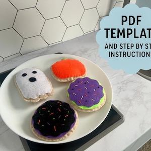 May include: A white plate holds four felt Halloween-themed treats: a ghost, a pumpkin, and two decorated cookies. The cookies are purple and black with colorful sprinkles. The image includes text that says "PDF TEMPLATE AND STEP BY STEP INSTRUCTIONS."