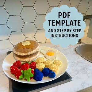 Puede incluir: Un juego de desayuno de fieltro en un plato blanco, que incluye panqueques, un huevo, fresas, arándanos y mantequilla. El plato está sobre una superficie negra. El texto en una forma de nube azul dice "PDF TEMPLATE AND STEP BY STEP INSTRUCTIONS."
